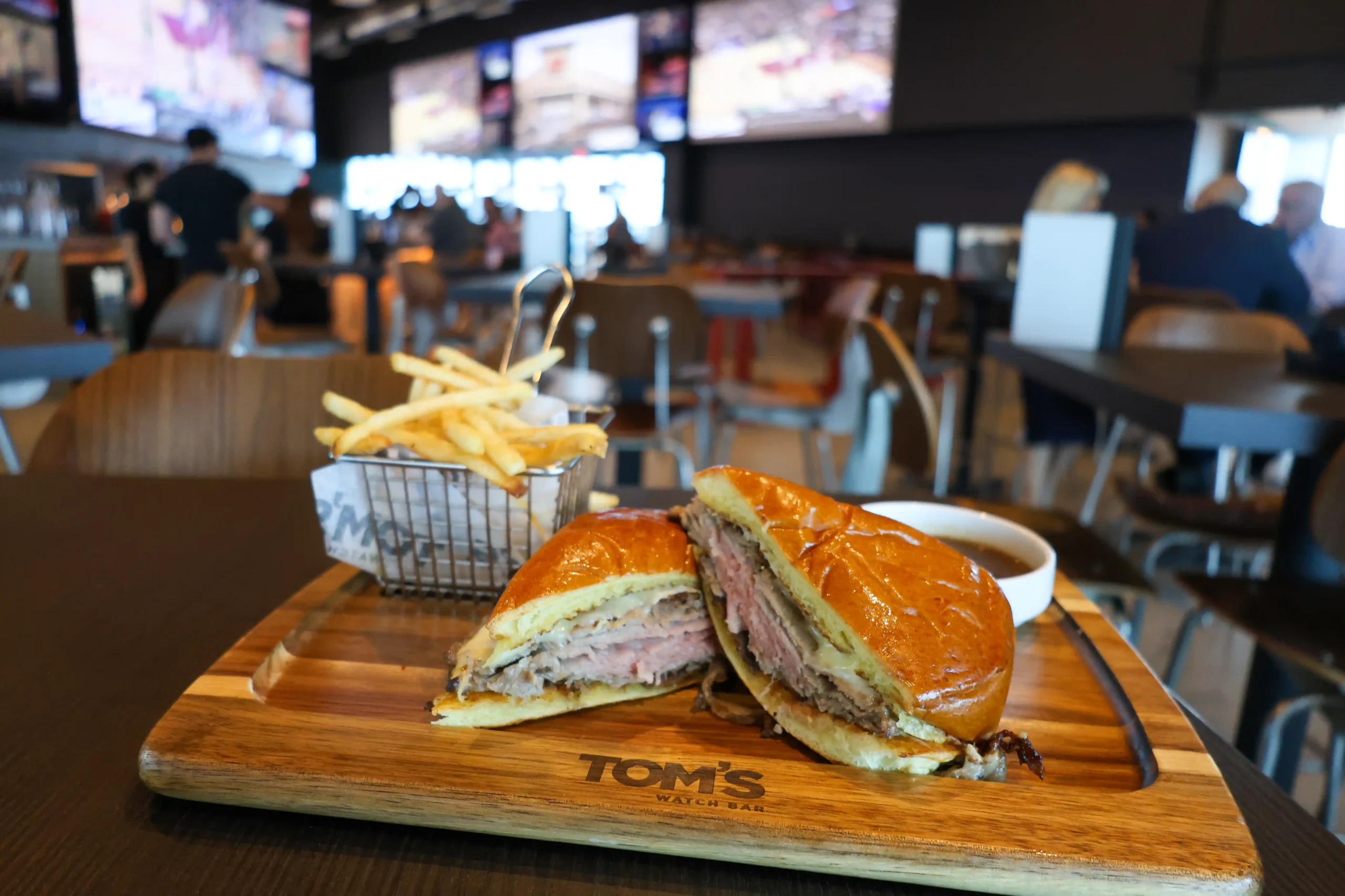 Sliced roast beef sandwich on a wooden board with fries in a small basket and dipping sauce, served inside a busy sports bar.