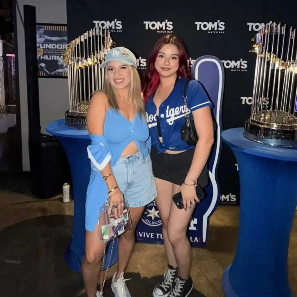 Pantone 294 Dodger Fans at Tom’s Watch Bar Two Dodgers fans in blue gear pose between World Series trophies in front of a Tom's Watch Bar backdrop.